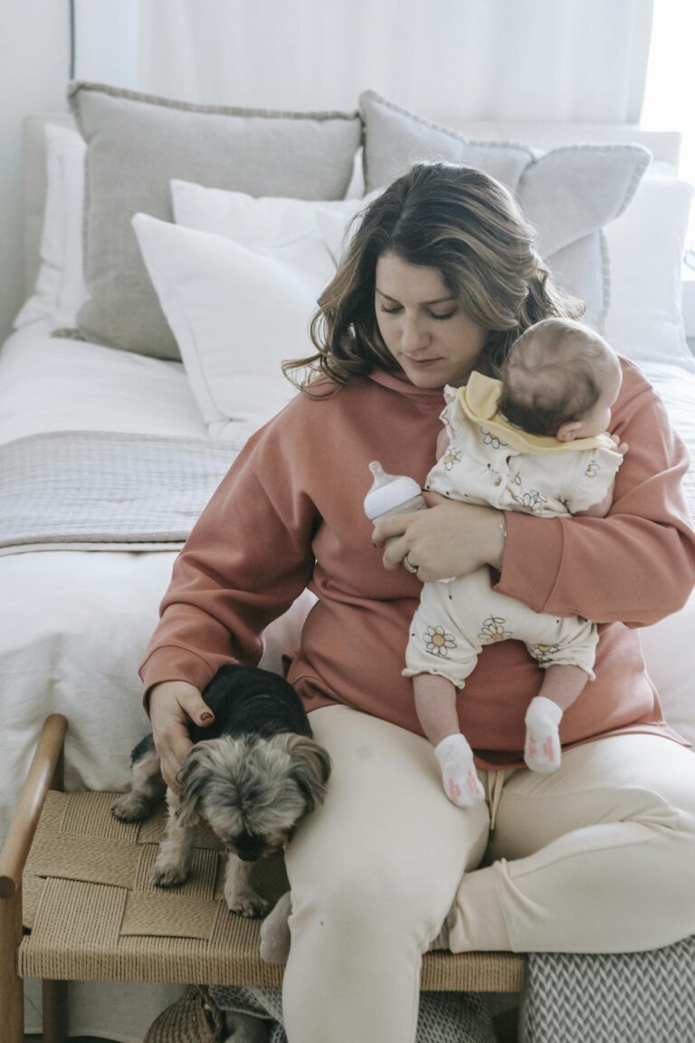 A nurturing mother holds her baby and caresses a small dog in a cozy bedroom setting.
